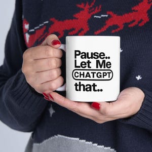 Puede incluir: Taza de cerámica blanca con el texto "Pause.. Let Me CHATGPT that.." en negro. La taza la sostiene una persona que lleva un jersey azul oscuro con un estampado de renos rojos y blancos.