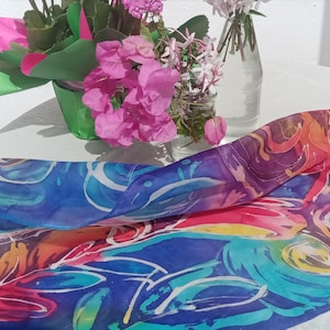 May include: A vibrant silk scarf featuring an abstract pattern in blue, purple, orange, and red hues. The scarf is displayed on a white surface, with flowers in the background. The design incorporates swirling forms and white details.