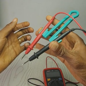 May include: A red and black multimeter probe set held together by a teal plastic organizer clip. The probes feature pointed metal tips and are connected to long electrical wires. A red digital multimeter with a screen and buttons is visible below.