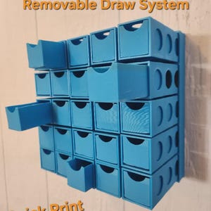 May include: A light blue modular storage system featuring twenty-five individual drawers arranged in a grid. The unit includes text that reads Fully Modular Removable Draw System, Quick Print, and 25 Grams Per Unit. Several drawers are pulled out.
