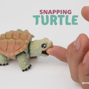 May include: A small, articulated snapping turtle figurine with a light green body and a textured, tan-colored shell. The turtle has its mouth open, appearing to bite a finger. The text SNAPPING TURTLE is printed above, with @FLEXI JIMGA below.