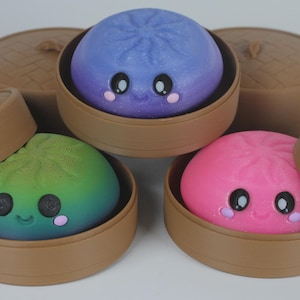 May include: Three round, smooth, colourful dumpling-shaped figures with smiling faces, large black eyes, and pink cheeks. Each figure is nestled inside a brown, circular, lidded bamboo-style steamer container. The dumplings are purple, green, and pink.