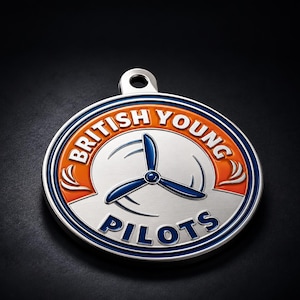 May include: A circular silver metal medallion featuring a blue three-blade propeller graphic in the center. The text BRITISH YOUNG PILOTS is embossed in white and blue lettering against an orange and silver background with a top hanging loop.