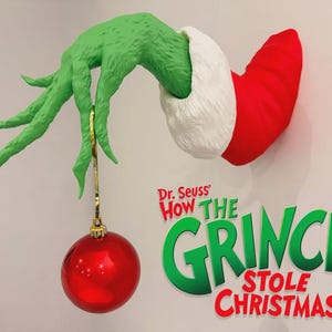 May include: A green textured hand emerging from a red and white cuff, holding a shiny red spherical ornament. Below the hand, the text reads Dr. Seuss How The Grinch Stole Christmas in green and red lettering against a plain light background.