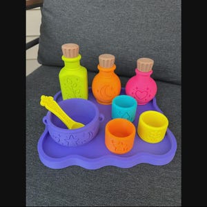 May include: A set of neon-coloured silicone play items including three bottles with cork-style stoppers, a cauldron with a matching spoon, and three small open cups, all arranged on a purple wavy-edged tray featuring embossed celestial and floral motifs.