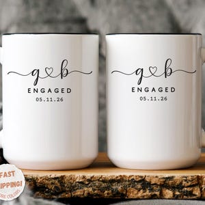May include: Two white ceramic mugs with black handles and rims. Each mug has the text "g&b ENGAGED 05.11.26" in black script. The mugs are displayed on a wooden surface.