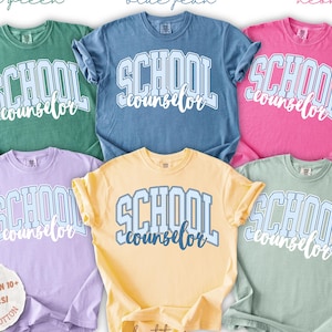 Puede incluir: Seis camisetas de manga corta en colores verde claro, azul vaquero, rosa ne&oacute;n, orqu&iacute;dea, mantequilla y laurel. Cada camiseta presenta el texto SCHOOL counselor en una fuente universitaria con un patr&oacute;n de rayas. Una etiqueta circular indica m&aacute;s de 10 colores.