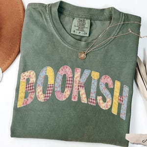 May include: A moss green t-shirt with the word "BOOKISH" in colorful patchwork letters. A gold necklace and a brown hat are next to the shirt. The shirt is made of 100% cotton.