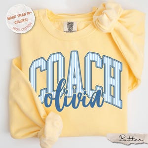 May include: A pale yellow long-sleeve sweatshirt featuring the word COACH in blue and white vertical stripes with the name olivia written below in a blue script font. The garment has a crew neckline and a Comfort Colors brand label inside.