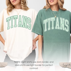 May include: Two short-sleeve crewneck t-shirts, one in ivory and one in light green, both featuring the word TITANS in a large, arched, striped collegiate font across the chest. The ivory shirt has dark green lettering, while the light green shirt has white lettering.