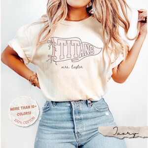May include: An ivory short-sleeve t-shirt featuring a graphic of a pennant flag with the text "TITANS" and "mrs. taylor" in a script font. The pennant includes bow details. A circular badge on the side states "MORE THAN 10+ COLORS!" and "100% COTTON".
