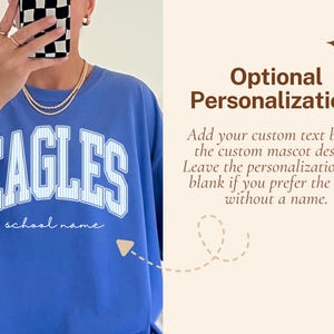 May include: A blue crewneck t-shirt featuring the word EAGLES in large white block letters with a thin blue pinstripe pattern. Below the word EAGLES, the text school name is printed in a white cursive script. The shirt has a relaxed fit.