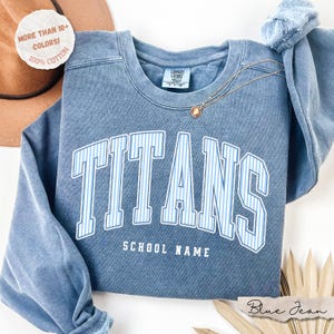 May include: A blue crewneck sweatshirt with a vintage wash finish. The front features the word TITANS in large, white-outlined letters with light blue vertical stripes, and the text SCHOOL NAME below. A gold necklace rests on the neckline.