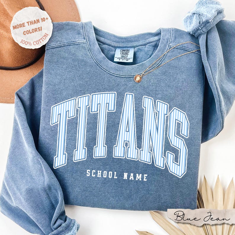 May include: A blue crewneck sweatshirt with a vintage wash finish. The front features the word TITANS in large, white-outlined letters with light blue vertical stripes, and the text SCHOOL NAME below. A gold necklace rests on the neckline.