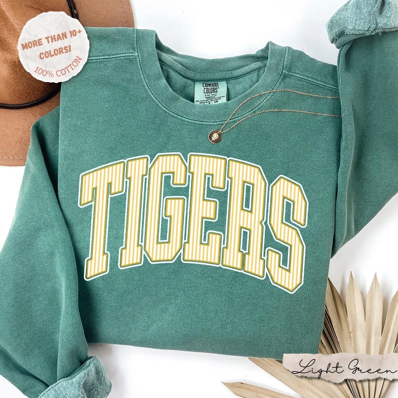 May include: A sage green crewneck sweatshirt featuring the word TIGERS in large, arched, yellow and white striped block lettering across the chest. The garment has a Comfort Colors label at the inner neckline and a thin gold-toned chain necklace.