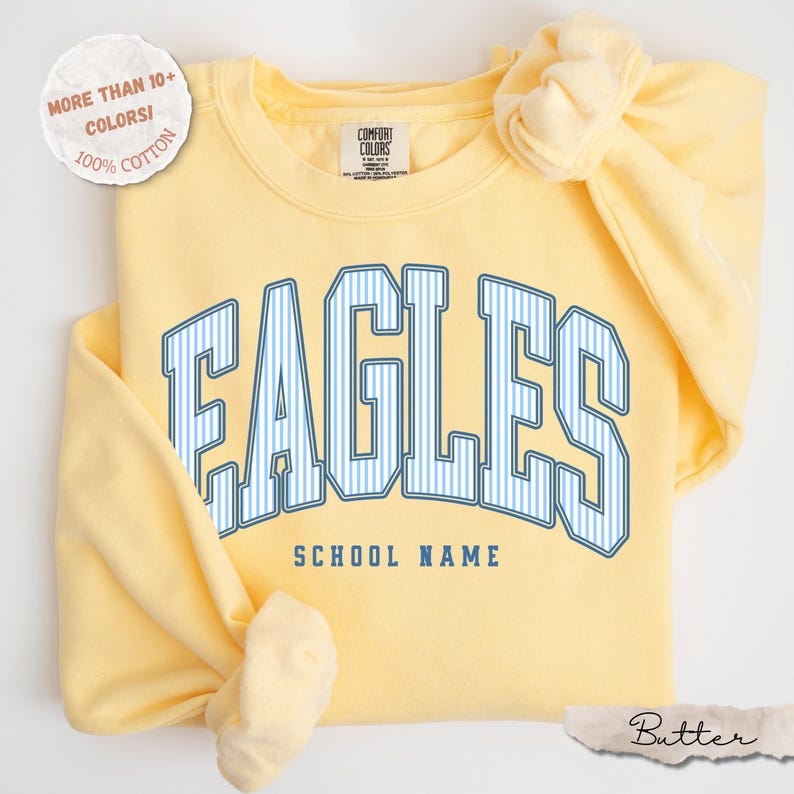 May include: A pale yellow crewneck sweatshirt featuring the word EAGLES in large, blue and white striped block letters with the text SCHOOL NAME printed below in blue. The garment label reads COMFORT COLORS and a circular badge states MORE THAN 10+ COLORS! 100% COTTON.