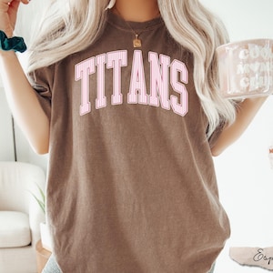 May include: A brown short-sleeve t-shirt featuring the word TITANS printed in a large, arched, pink and white striped collegiate font across the chest. The shirt has a crew neckline and a relaxed fit, showcasing a solid, muted brown fabric color.