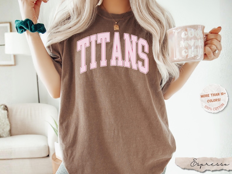 May include: A brown short-sleeve t-shirt featuring the word TITANS printed in a large, arched, pink and white striped collegiate font across the chest. The shirt has a crew neckline and a relaxed fit, showcasing a solid, muted brown fabric color.