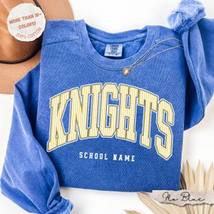 May include: A blue crewneck sweatshirt featuring the word KNIGHTS in large yellow striped lettering with the text SCHOOL NAME underneath. The garment has a Comfort Colors label at the inner neckline and a thin gold chain necklace resting on top.