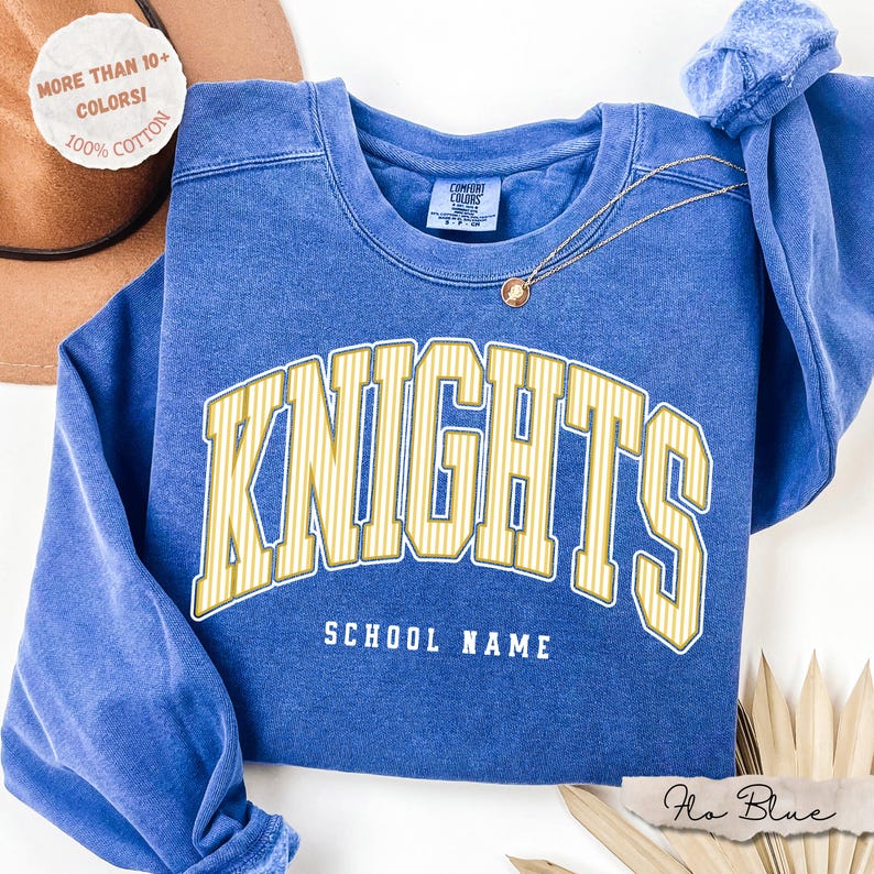 May include: A blue crewneck sweatshirt featuring the word KNIGHTS in large yellow striped lettering with the text SCHOOL NAME underneath. The garment has a Comfort Colors label at the inner neckline and a thin gold chain necklace resting on top.