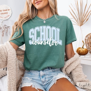 Puede incluir: Una camiseta de manga corta color verde salvia que presenta la palabra SCHOOL en una fuente universitaria grande de color blanco con rayas azules, con la palabra counselor escrita debajo en una letra cursiva blanca. Tiene cuello redondo y corte holgado.