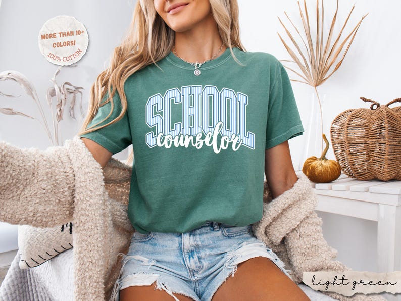 Puede incluir: Una camiseta de manga corta color verde salvia que presenta la palabra SCHOOL en una fuente universitaria grande de color blanco con rayas azules, con la palabra counselor escrita debajo en una letra cursiva blanca. Tiene cuello redondo y corte holgado.
