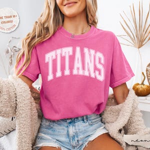 May include: A bright pink short-sleeved t-shirt featuring the word TITANS in a white, arched, pinstriped collegiate font across the chest. The shirt has a crew neckline and a relaxed fit, paired with light blue denim shorts and a fuzzy cardigan.