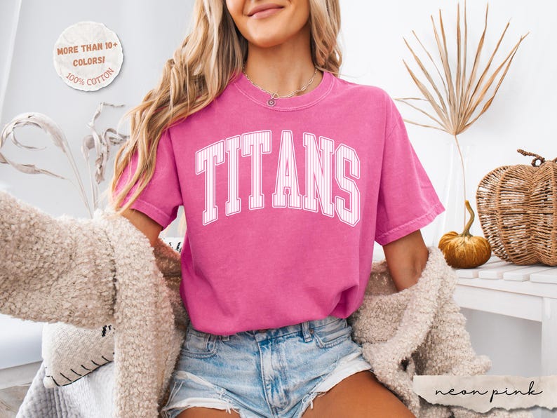 May include: A bright pink short-sleeved t-shirt featuring the word TITANS in a white, arched, pinstriped collegiate font across the chest. The shirt has a crew neckline and a relaxed fit, paired with light blue denim shorts and a fuzzy cardigan.