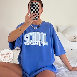 Puede incluir: Una camiseta azul b&iacute;garo con el texto SCHOOL counselor en letras blancas y azul claro, combinando estilo bloque y cursiva. La prenda incluye un gr&aacute;fico circular que dice MORE THAN 10+ COLORS! 100% COTTON y una etiqueta Flo blue.