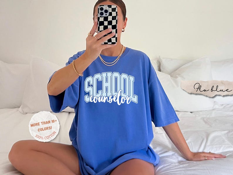 Puede incluir: Una camiseta azul b&iacute;garo con el texto SCHOOL counselor en letras blancas y azul claro, combinando estilo bloque y cursiva. La prenda incluye un gr&aacute;fico circular que dice MORE THAN 10+ COLORS! 100% COTTON y una etiqueta Flo blue.