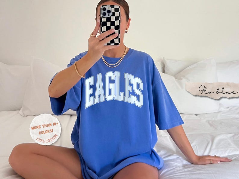 May include: A periwinkle blue short-sleeved t-shirt featuring the word EAGLES in large white block letters with a thin pinstripe pattern. A circular white graphic on the left reads MORE THAN 10+ COLORS! 100% COTTON. A script text reads Flo blue.