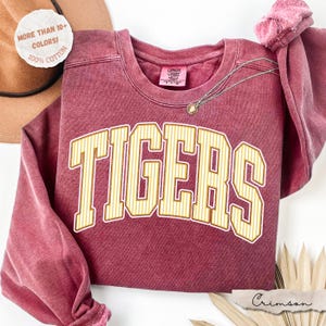 May include: A crimson red crewneck sweatshirt with a vintage-style wash. The front features the word TIGERS in large, arched, yellow and white striped block letters. The interior neck label reads Comfort Colors, and the fabric is 100% cotton.