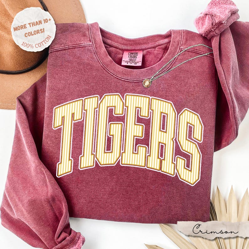 May include: A crimson red crewneck sweatshirt with a vintage-style wash. The front features the word TIGERS in large, arched, yellow and white striped block letters. The interior neck label reads Comfort Colors, and the fabric is 100% cotton.