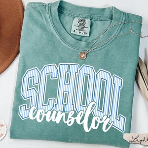 Puede incluir: Una camiseta de manga corta color verde salvia con cuello redondo. La prenda presenta la palabra SCHOOL en letras may&uacute;sculas grandes a rayas azules y blancas, con la palabra counselor escrita debajo en letra cursiva blanca. Un collar descansa encima.