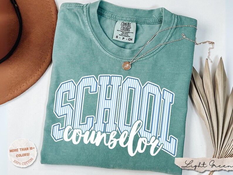 Puede incluir: Una camiseta de manga corta color verde salvia con cuello redondo. La prenda presenta la palabra SCHOOL en letras may&uacute;sculas grandes a rayas azules y blancas, con la palabra counselor escrita debajo en letra cursiva blanca. Un collar descansa encima.