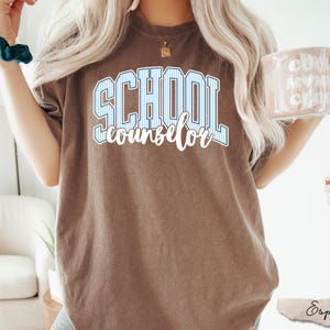 Puede incluir: Una camiseta marr&oacute;n de manga corta con el texto SCHOOL en letras de bloque a rayas azules y blancas, con la palabra counselor escrita debajo en letra cursiva blanca. La prenda tiene cuello redondo y un corte relajado de estilo casual.