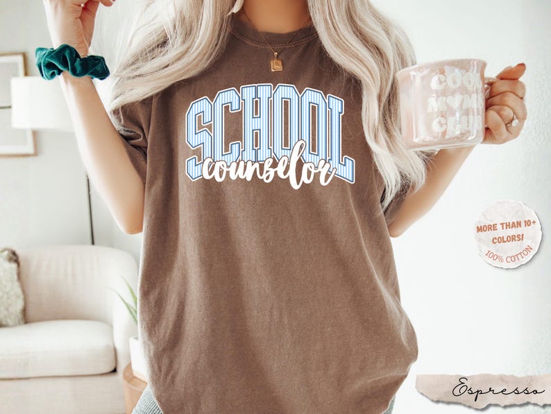 Puede incluir: Una camiseta marr&oacute;n de manga corta con el texto SCHOOL en letras de bloque a rayas azules y blancas, con la palabra counselor escrita debajo en letra cursiva blanca. La prenda tiene cuello redondo y un corte relajado de estilo casual.