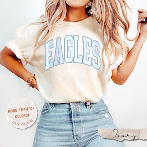 May include: An ivory short-sleeve t-shirt featuring the word EAGLES in a large, arched, light blue and white pinstriped collegiate font. The shirt is paired with light blue denim jeans. A circular graphic in the corner reads MORE THAN 10+ COLORS! 100% COTTON.