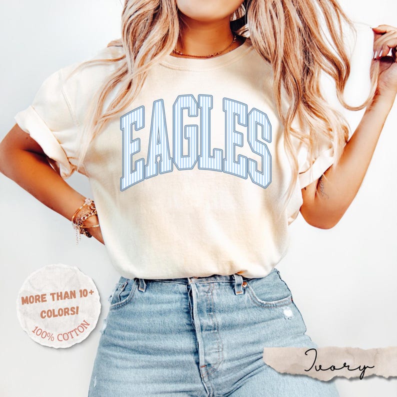 May include: An ivory short-sleeve t-shirt featuring the word EAGLES in a large, arched, light blue and white pinstriped collegiate font. The shirt is paired with light blue denim jeans. A circular graphic in the corner reads MORE THAN 10+ COLORS! 100% COTTON.