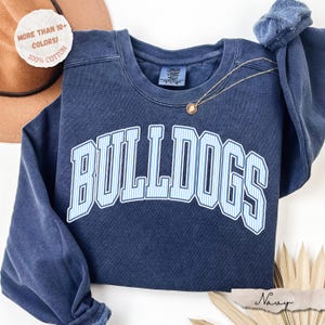 May include: A navy blue crewneck sweatshirt featuring the word BULLDOGS in a white and light blue vertical striped font with a white outline. The garment has a Comfort Colors label at the inner neckline and a thin gold chain necklace resting on top.