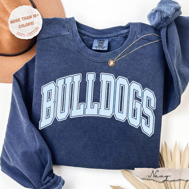 May include: A navy blue crewneck sweatshirt featuring the word BULLDOGS in a white and light blue vertical striped font with a white outline. The garment has a Comfort Colors label at the inner neckline and a thin gold chain necklace resting on top.