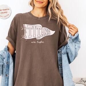 May include: An espresso brown short-sleeve t-shirt featuring a white pennant graphic with a light pink checkered pattern. The text "TITANS" is printed in large white block letters inside the pennant, with "mrs. taylor" written below in script.