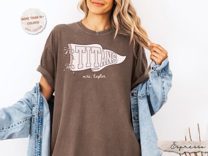 May include: An espresso brown short-sleeve t-shirt featuring a white pennant graphic with a light pink checkered pattern. The text "TITANS" is printed in large white block letters inside the pennant, with "mrs. taylor" written below in script.