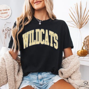 May include: A black short-sleeve t-shirt featuring the word WILDCATS in a yellow and white arched varsity-style font. The shirt includes a circular graphic in the top left corner with the text MORE THAN 10+ COLORS! 100% COTTON.