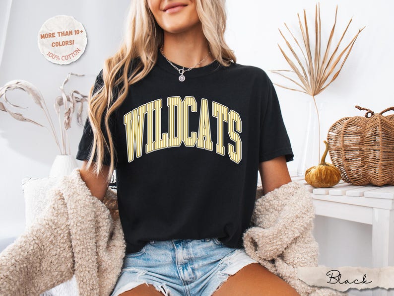 May include: A black short-sleeve t-shirt featuring the word WILDCATS in a yellow and white arched varsity-style font. The shirt includes a circular graphic in the top left corner with the text MORE THAN 10+ COLORS! 100% COTTON.