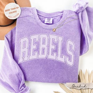 May include: A lavender long-sleeve crewneck sweatshirt with a vintage-wash finish. The front features the word REBELS in large, arched, white-outlined block letters with vertical pinstripes. A small Comfort Colors brand tag is inside the collar.