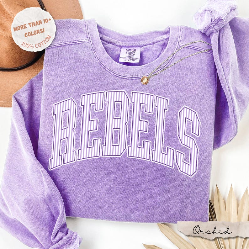 May include: A lavender long-sleeve crewneck sweatshirt with a vintage-wash finish. The front features the word REBELS in large, arched, white-outlined block letters with vertical pinstripes. A small Comfort Colors brand tag is inside the collar.
