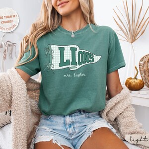 May include: A green short-sleeve t-shirt featuring a white and green checkered pennant graphic with the word LIONS in dark green block letters and the text mrs. taylor in white script. A circular badge states MORE THAN 10+ COLORS! 100% COTTON.