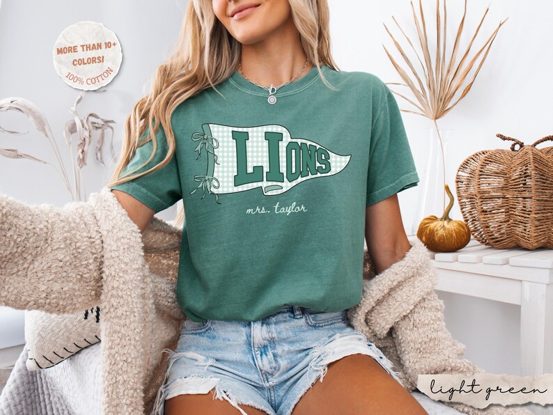 May include: A green short-sleeve t-shirt featuring a white and green checkered pennant graphic with the word LIONS in dark green block letters and the text mrs. taylor in white script. A circular badge states MORE THAN 10+ COLORS! 100% COTTON.