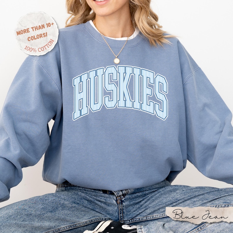 May include: A dusty blue crewneck sweatshirt featuring the word HUSKIES in a white and blue striped collegiate font across the chest. A circular badge in the upper left corner reads MORE THAN 10+ COLORS! 100% COTTON. The garment is worn with blue jeans.